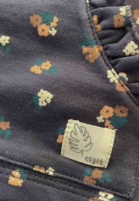 Tkanina s cvjetnim uzorkom u tamnosivoj boji, s malim cvjetovima u narančastoj, žutoj i zelenoj boji. Uključuje etiketu krem boje s detaljima logotipa.
