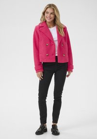 Veste cropped double boutonnage fuchsia en tissu texturé, avec des boutons dorés et des revers crantés, assortie à un pantalon noir ajusté et des chaussures.