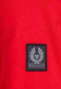 Tessuto rosso caratterizzato da una superficie liscia, con un'etichetta rettangolare nera in evidenza che mostra un logo e la parola "BELSTAFF" in grigio.