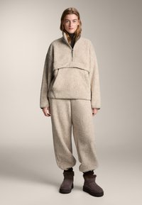 Bež pulover iz flisa s polovičnim zadrgo, večplastno zasnovo in usklajenimi hlačami. Teksturirana tkanina, ohlapen kroj, rjavi gležnarji z obrobom iz krzna.