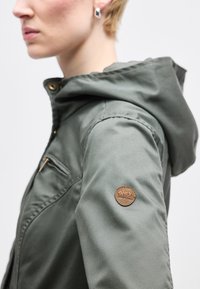 Άτομο που φοράει γκρι μπουφάν με κουκούλα, με καφέ στρογγυλό κέντημα που γράφει "ONLY" στο πάνω μέρος του μανικιού και ασημένιο κρίκο στα αυτιά.