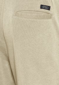 Beiger Pullover mit weich gestrickter Textur, rechteckiger Rückentasche und einem Etikett mit weißem Text auf dunklem Hintergrund.