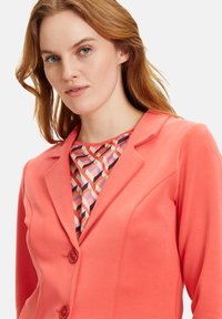 Korallenblazer mit zwei Knöpfen, taillierter Passform und Reverskragen, kombiniert mit einem geometrisch gemusterten Innenoberteil in Pastellfarben.