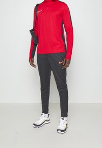 Rode Nike longsleeve top met zwarte zijaccenten, gecombineerd met donkergrijze joggingbroek met een oranje Nike-logo. Witte en zwarte Nike sneakers.