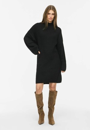 Woman wearing a black long-sleeve knit dress with high brown suede boots, standing with one hand on hip against a plain white background.