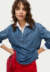 Blauwe denim shirt met een placket-halslijn, twee borstzakken en lange mouwen. Draagt over een witte shirt met kraag. Rode handtas zichtbaar.