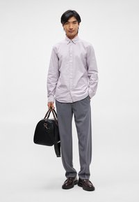 Randig ljuslila skjorta, grå byxor, glänsande bruna skor. Håller en svart läderväska med kontrasterande bruna handtag.