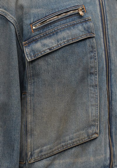 Denim jacket featuring a light blue color, prominent stitching, a zippered chest pocket, and a large flap pocket for utility.