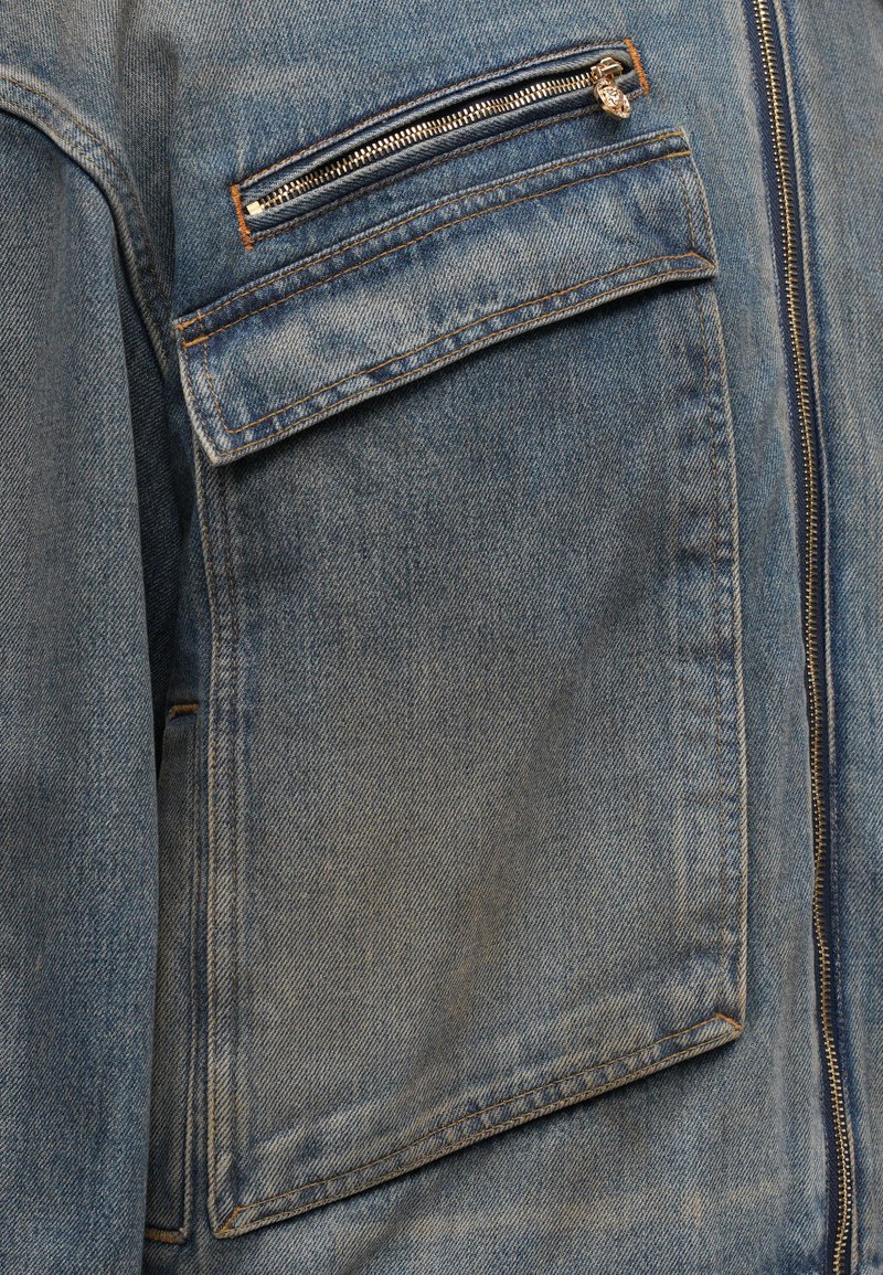 Denimjacke in hellblauer Farbe, mit auffälliger Naht, einer Reißverschlusstasche auf der Brust und einer großen Klapptasche für praktischen Einsatz.