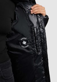 Chaqueta de plumas negra con una textura acolchada brillante. El logo interior dice "KHUJO ORIGINAL." Incluye bolsillos laterales y un diseño elegante.