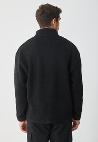 Giacca in pile nera con superficie texturizzata, colletto alto e maniche lunghe. Il retro presenta un design semplice, senza loghi o accenti visibili.