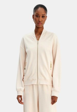 Donna che indossa una giacca beige chiaro con zip, con polsini a coste e dettagli in oro sulle tasche laterali, in piedi davanti a uno sfondo semplice.