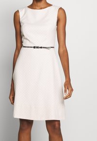 Un vestido sin mangas de color rosa claro que presenta un sutil patrón de diamantes, cintura definida y un delgado cinturón plateado que acentúa la cintura.