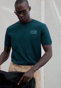 Tirkīzzils īsroku T-krekls no mīksta kokvilnas ar kvadrātveida, teksturētu logotipa uzšuvumu uz krūtīm. Valkāts kopā ar haki krāsas biksēm, izceļot ikdienišķu stilu.