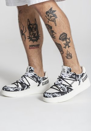 Nohy s čiernymi tetovaniami vrátane psa a anjela, obuté v bielych a čiernych teniskách s vzorom s nápisom "GIANNI" a čiernymi šnúrkami.