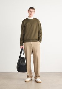 Sweatshirt vert olive avec des poignets côtelés, associé à un pantalon beige plissé et des baskets blanches. Un sac noir est tenu dans une main.