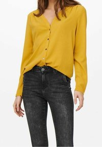 Blouse jaune à manches longues avec un col en V, des boutons sur le devant et une texture lisse, associée à un jean slim gris foncé avec un lavage subtil.