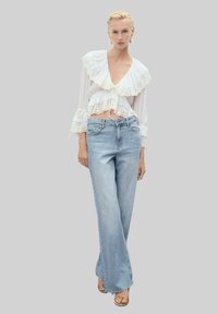 Vit genomskinlig blus med volangkanter och trumpetärmar; ihop med ljusblå vida jeans. Outfiten har en avslappnad, lager-på-lager design.