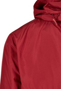 Červená vnější bunda s hladkým, strukturovaným povrchem, nastavitelnou kapucí se šňůrkami a uvolněným střihem.