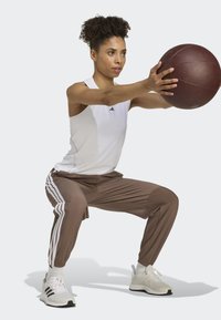 White tank top and brown athletic pants with white stripes, performing a squat while holding a brown medicine ball. White sneakers with accents.