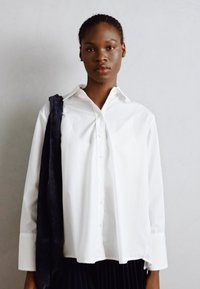 Camicia bianca con maniche lunghe, caratterizzata da un colletto classico e una vestibilità ampia. Tessuto scuro appoggiato su una spalla. Texture liscia.