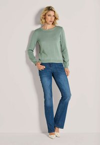 Frau mit schulterlangem blonden Haar, die einen hellgrünen Pullover, bluejeans und cremefarbene spitze Schuhe trägt, steht vor einer schlichten Wand.