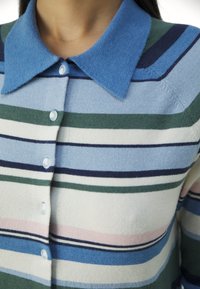 Gros plan d’un cardigan tricoté à rayures avec col bleu et boutons blancs, présentant des bandes horizontales en bleu, vert, marine, rose et crème.