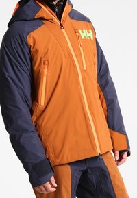 Veste orange et marine avec une capuche, comprenant deux poches latérales zippées, des poignets en Velcro et un logo vif. Fabriquée en matériau résistant aux intempéries.