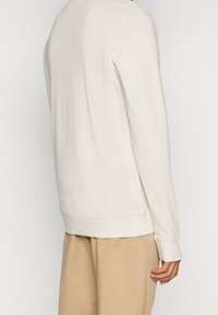 Persona vestida con un sudadera de manga larga color crema y pantalones beige, vista desde atrás con un brazo relajado a su lado.