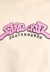 Felpa di un beige chiaro con una grafica ricamata in peluche rosa e nera "Santa Cruz Skateboards". Presenta un collo rotondo e una texture morbida.