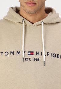 Beige hoodie with a front logo featuring "TOMMY HILFIGER EST. 1985." Cotton blend fabric, drawstrings with metallic tips, and embroidered details.