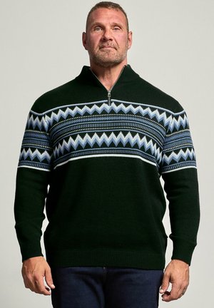 Hombre con cabello corto y barba que lleva un suéter negro con patrones en zigzag azules y blancos y un cuello con media cremallera, de pie frente a un fondo liso.