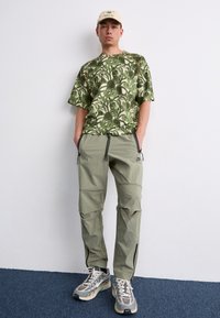 Green floral patterned oversized shirt, light green cargo pants with pockets, beige cap, and multicolored sneakers with a chunky sole.
