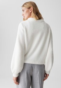 Pullover bianco e morbido con una consistenza soffice e scollatura rotonda; indossato con pantaloni grigi su misura, caratterizzati da maniche ampie a palloncino.