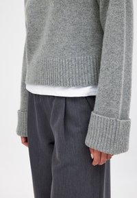 Pelēka adīta džemperis ar ribinātām piedurknēm, uzvilkts virs baltās krekla, apvienots ar tumši pelēkām plecēm biksēm. Mīksta tekstūra, ikdienas piegriezums.