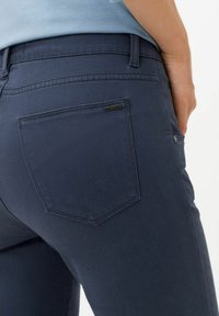 Jeans en denim bleu foncé avec une poche arrière en forme triangulaire et un petit logo métallique, affichant une texture lisse et soyeuse.