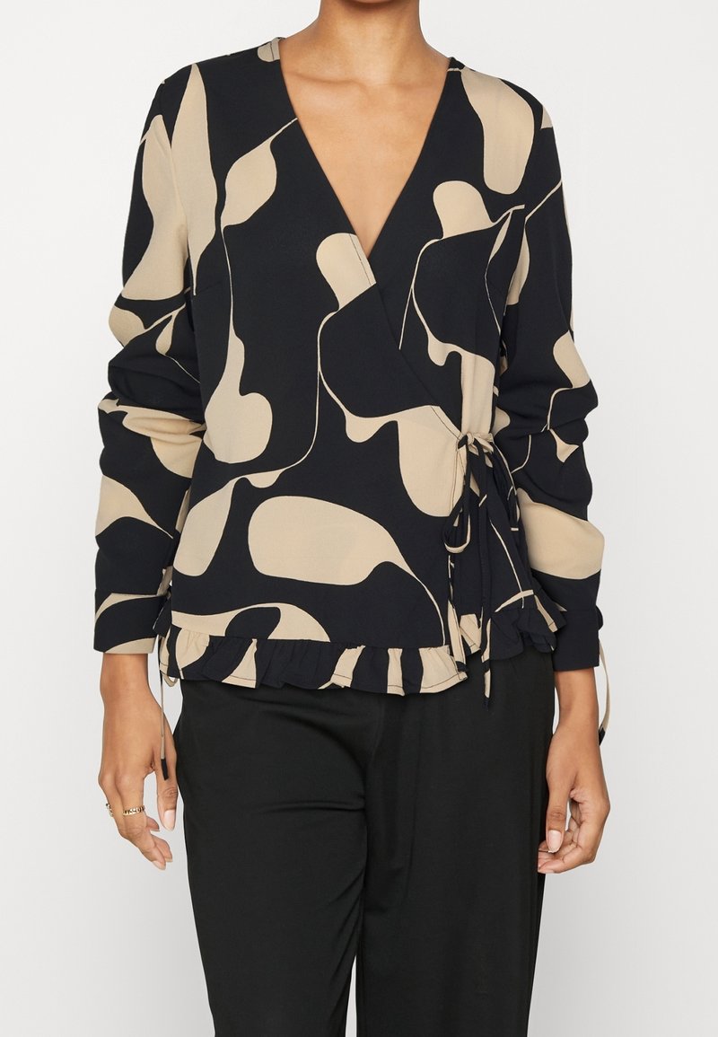 Femme portant un chemisier cache-cœur à motif abstrait noir et beige avec manches longues et ourlet volanté, associé à un pantalon noir.