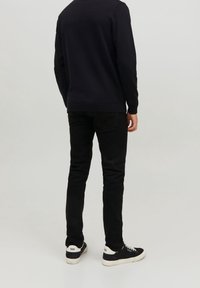 Schwarze Jeans, schmale Passform, aus Baumwolle, kombiniert mit einem dunklen Pullover. Schwarze Sneakers mit weißen Akzenten runden das Outfit ab, von hinten betrachtet.