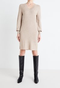 Beige stickad klänning med V-ringning och långa ärmar, med rak silhuett. Matchas med eleganta svarta knähöga stövlar.