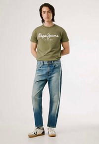 Oliwkowa bawełniana koszulka z napisem "Pepe Jeans London", zestawiona z jasnoniebieskimi, wyblakłymi jeansami o prostych nogawkach oraz białymi trampkami z czarnymi akcentami.