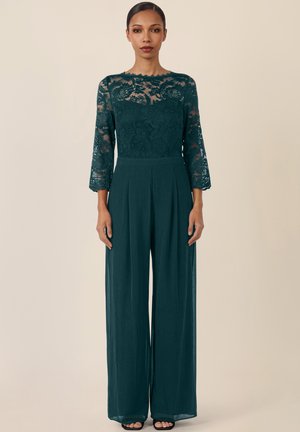 Frau steht und trägt ein dunkelgrünes Spitzenoberteil mit dreiviertel Ärmeln und weiten Hosen vor einem einfarbigen beigen Hintergrund.