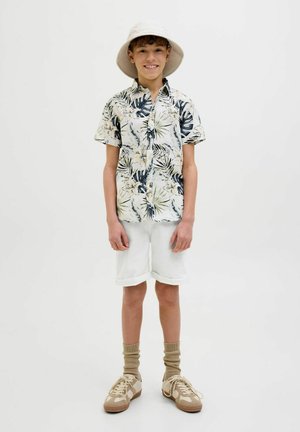 Chemise à manches courtes avec un motif de feuilles tropicales dans des tons de vert et de crème, associée à un short blanc enroulé, des baskets beiges et un léger chapeau de pêcheur.