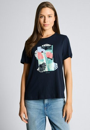 Kobieta o długich brązowych włosach ubrana w granatowy T-shirt z kolorowym, abstrakcyjnym wzorem kwiatowym w kształcie wachlarza oraz jasnoniebieskie jeansy, stojąca na szarym tle.