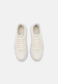 Beige lädersneakers med vita detaljer, rund tå, perforerat mönster och platta snören. Innehåller "Nike Air"-logotyp på tungan.