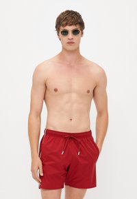 Vörös fürdőshorts könnyű anyagból, rugalmas derékkal és húzózsinórral, oldalsó zsebekkel és kis fehér díszítőelemekkel.