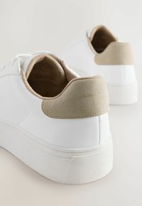 Baskets blanches avec une tige en cuir lisse, un accent au niveau du talon en suède gris, une épaisse semelle en caoutchouc blanc avec un grip texturé et une doublure beige.
