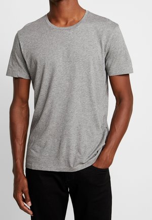 Homme portant un t-shirt gris uni à manches courtes, une main dans la poche et un pantalon noir sur fond blanc.