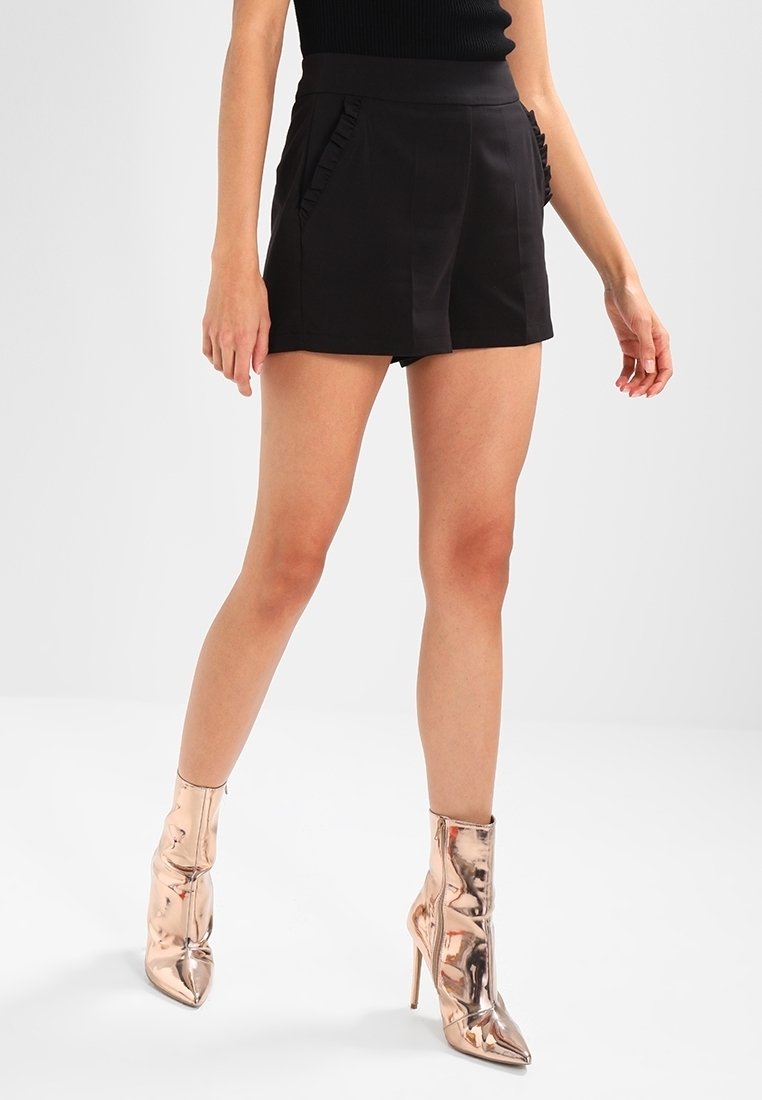 Shorts noirs taille haute avec poches à volants, présentant un design ajusté et un tissu lisse. Associés à des bottines métalliques brillantes.