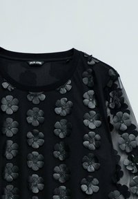 Top noir avec des appliqués floraux texturés dans des teintes sombres, des manches transparentes et un col rond. Fabriqué à partir d'un mélange de matériaux.