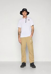 Wit shirt met korte mouwen en een ritssluiting bij de kraag, beige cargobroek met zijzakken, zwarte bucket hat en zwarte wandelschoenen.